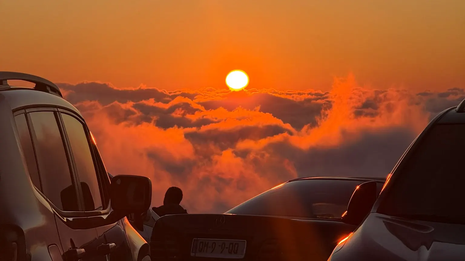 The sun setting into the clouds at Gomismta, seen between parked cars